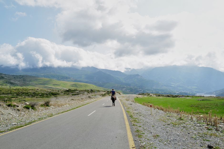 A person riding a horse down an open road surrounded by lush mountains and cloudy skies.